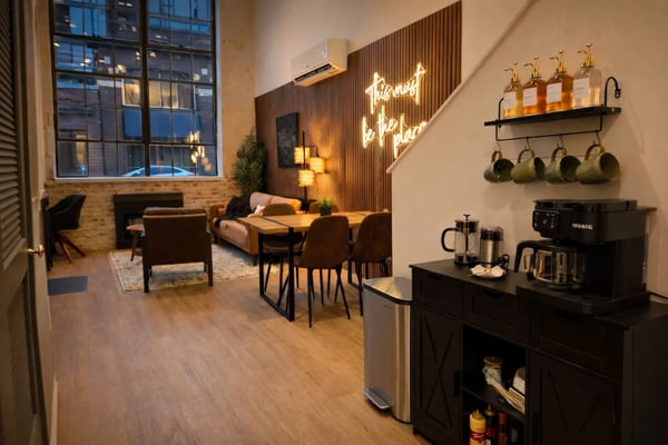 Modern loft kitchen and lounge with neon sign, dining table, coffee station, and large windows overlooking city at dusk